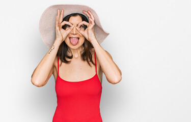 Beautiful brunette woman wearing swimsuit and summer hat doing ok gesture like binoculars sticking tongue out, eyes looking through fingers. crazy expression.