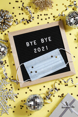 black letter board with text BYE BYE 2021 with decoration and mask on yellow background.