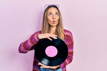 Beautiful hispanic woman wearing hippie style holding vinyl disc making fish face with mouth and...