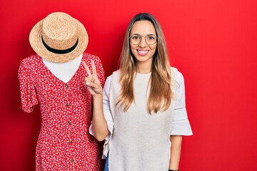 Beautiful hispanic woman standing by manikin smiling with happy face winking at the camera doing...