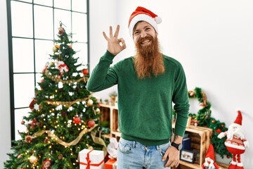 Redhead man with long beard wearing christmas hat by christmas tree smiling positive doing ok sign with hand and fingers. successful expression.