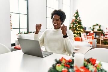 Young african american man using laptop sitting on the table by christmas tree excited for success...