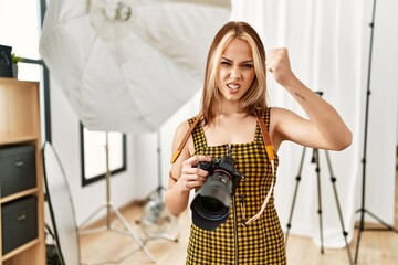 Young caucasian photographer girl holding professional camera at photography studio annoyed and frustrated shouting with anger, yelling crazy with anger and hand raised