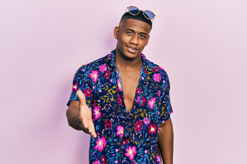 Young black man wearing hawaiian shirt and sunglasses smiling friendly offering handshake as greeting and welcoming. successful business.
