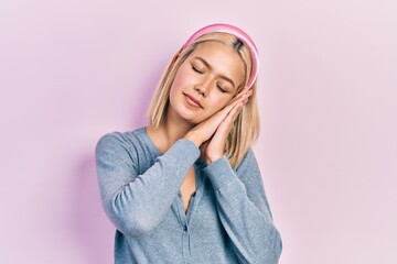 Beautiful blonde woman standing over pink background sleeping tired dreaming and posing with hands together while smiling with closed eyes.