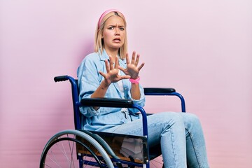 Beautiful blonde woman sitting on wheelchair afraid and terrified with fear expression stop gesture...