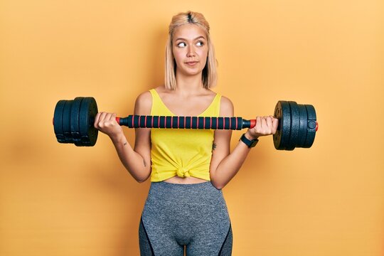 Beautiful blonde woman wearing sportswear using dumbbells smiling looking to the side and staring away thinking.