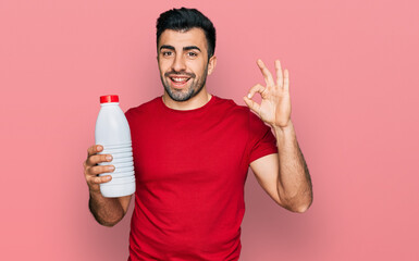 Hispanic man with beard holding liter bottle of milk doing ok sign with fingers, smiling friendly gesturing excellent symbol