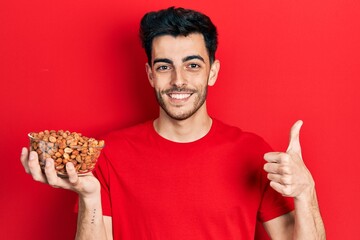 Young hispanic man holding peanuts smiling happy and positive, thumb up doing excellent and approval sign