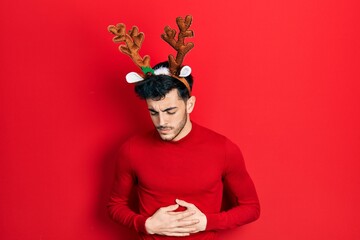 Young hispanic man wearing cute christmas reindeer horns with hand on stomach because indigestion,...