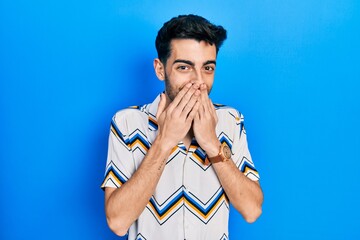 Young hispanic man wearing casual clothes laughing and embarrassed giggle covering mouth with...