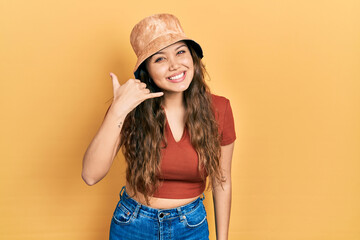 Young hispanic girl wearing casual clothes and hat smiling doing phone gesture with hand and...