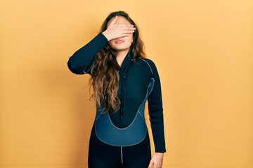 Young hispanic girl wearing diver neoprene uniform covering eyes with hand, looking serious and...