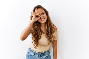 Beautiful hispanic woman standing over isolated while background doing ok gesture with hand smiling, eye looking through fingers with happy face.