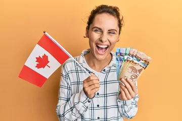 Papier Peint photo Canada Young brunette woman holding canada flag and dollars smiling and laughing hard out loud because funny crazy joke.