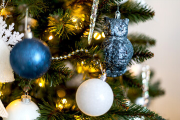 Blue and white balls and an owl hang on a christmas tree with glowing orange lights. New year concept, close up side view