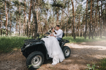 Stylish young groom in a shirt and a beautiful bride in a white long dress are sitting embracing on a black ATV in the forest. Wedding photography, portrait.