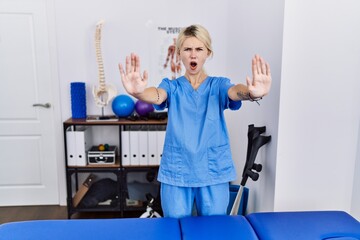 Young physiotherapist woman working at pain recovery clinic doing stop gesture with hands palms, angry and frustration expression