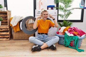 Young blonde woman doing laundry sitting by washing machine smiling pointing to head with both hands finger, great idea or thought, good memory