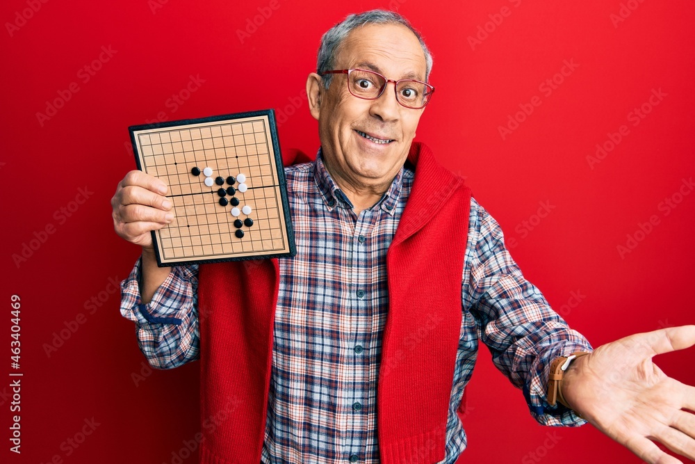 Poster handsome senior man with grey hair holding asian go game board celebrating achievement with happy sm