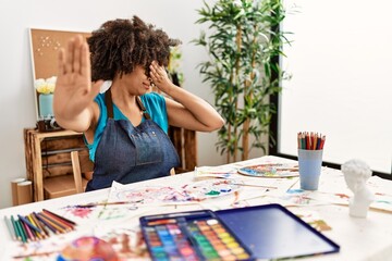 Beautiful african american woman with afro hair painting at art studio covering eyes with hands and doing stop gesture with sad and fear expression. embarrassed and negative concept.