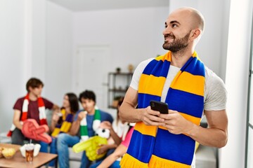 Group of young hispanic friends watching and supporting soccer match. Man smiling happy and using smartphone at home.