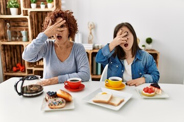 Family of mother and down syndrome daughter sitting at home eating breakfast peeking in shock covering face and eyes with hand, looking through fingers with embarrassed expression.