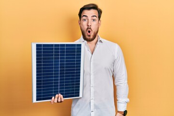 Handsome caucasian man with beard holding photovoltaic solar panel scared and amazed with open...