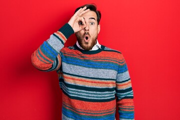 Handsome caucasian man with beard wearing elegant wool winter sweater doing ok gesture shocked with surprised face, eye looking through fingers. unbelieving expression.