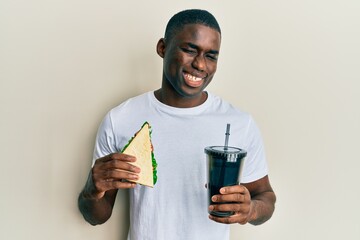 Young african american man eating sandwich and drinking soda winking looking at the camera with...