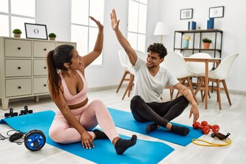 Young latin couple smiling happy training at home. Sitting on the floor high five.