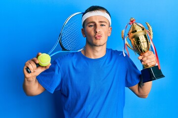 Young caucasian man playing tennis holding trophy looking at the camera blowing a kiss being lovely...