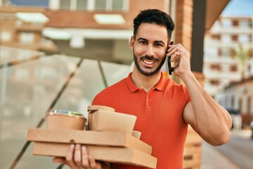 Young hispanic man talking on the smartphone and holding take away food at the city