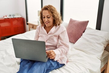 Middle age caucasian woman using laptop sitting on the bed at bedroom.