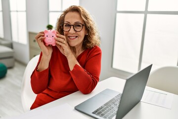 Middle age caucasian woman holding piggy bank sitting on the table at home.