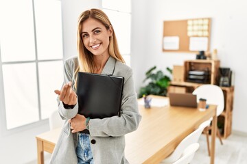 Blonde business woman at the office beckoning come here gesture with hand inviting welcoming happy and smiling