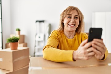 Middle age blonde woman smiling happy using smartphone at new home.