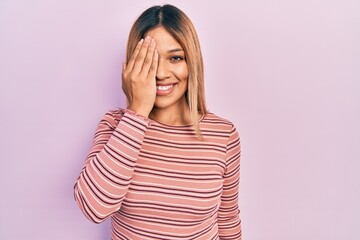 Beautiful hispanic woman wearing casual striped sweater covering one eye with hand, confident smile on face and surprise emotion.