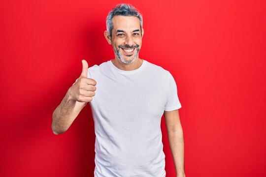 Handsome middle age man with grey hair wearing casual white t shirt smiling happy and positive, thumb up doing excellent and approval sign