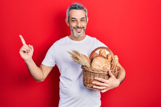 Handsome middle age man with grey hair holding wicker basket with bread smiling happy pointing with hand and finger to the side