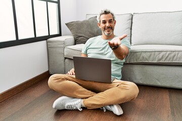 Middle age hispanic man using laptop sitting on the floor at the living room smiling cheerful offering palm hand giving assistance and acceptance.