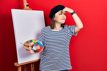 Middle age hispanic woman standing drawing with palette by painter easel stand tired rubbing nose...