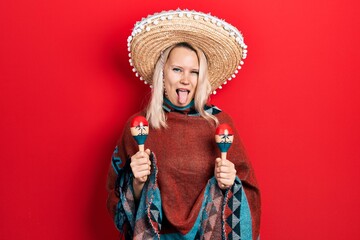 Beautiful caucasian blonde woman wearing festive mexican poncho and maracas sticking tongue out happy with funny expression.