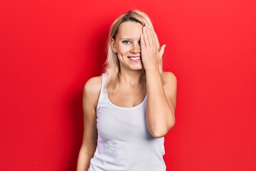 Beautiful caucasian blonde woman wearing casual white t shirt covering one eye with hand, confident smile on face and surprise emotion.