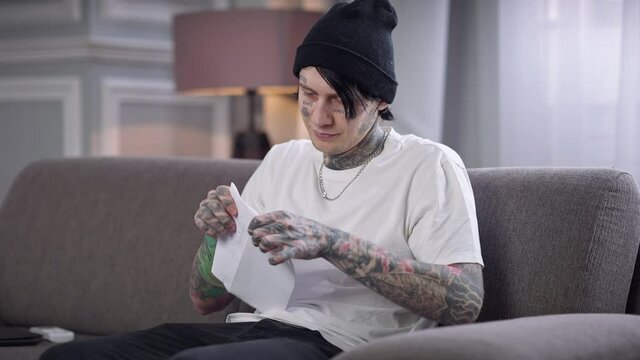 Positive tattooed man in casual clothes and hat putting letter in envelope. Portrait of confident Caucasian guy sitting on couch in living room packing preparing mail to send