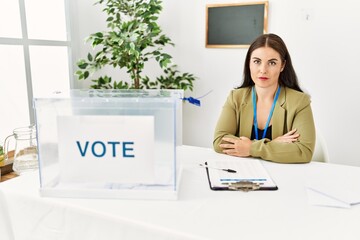Young brunette woman sitting at election table with voting ballot relaxed with serious expression...