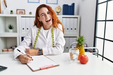 Young redhead woman nutritionist doctor at the clinic winking looking at the camera with sexy...