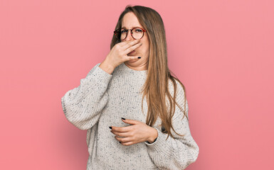 Young blonde woman wearing casual sweater and glasses smelling something stinky and disgusting,...