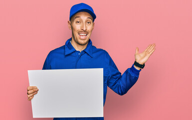 Bald man with beard wearing builder jumpsuit uniform holding empty banner celebrating victory with happy smile and winner expression with raised hands