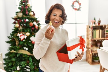 Middle age hispanic woman holding gift standing by christmas tree at home.